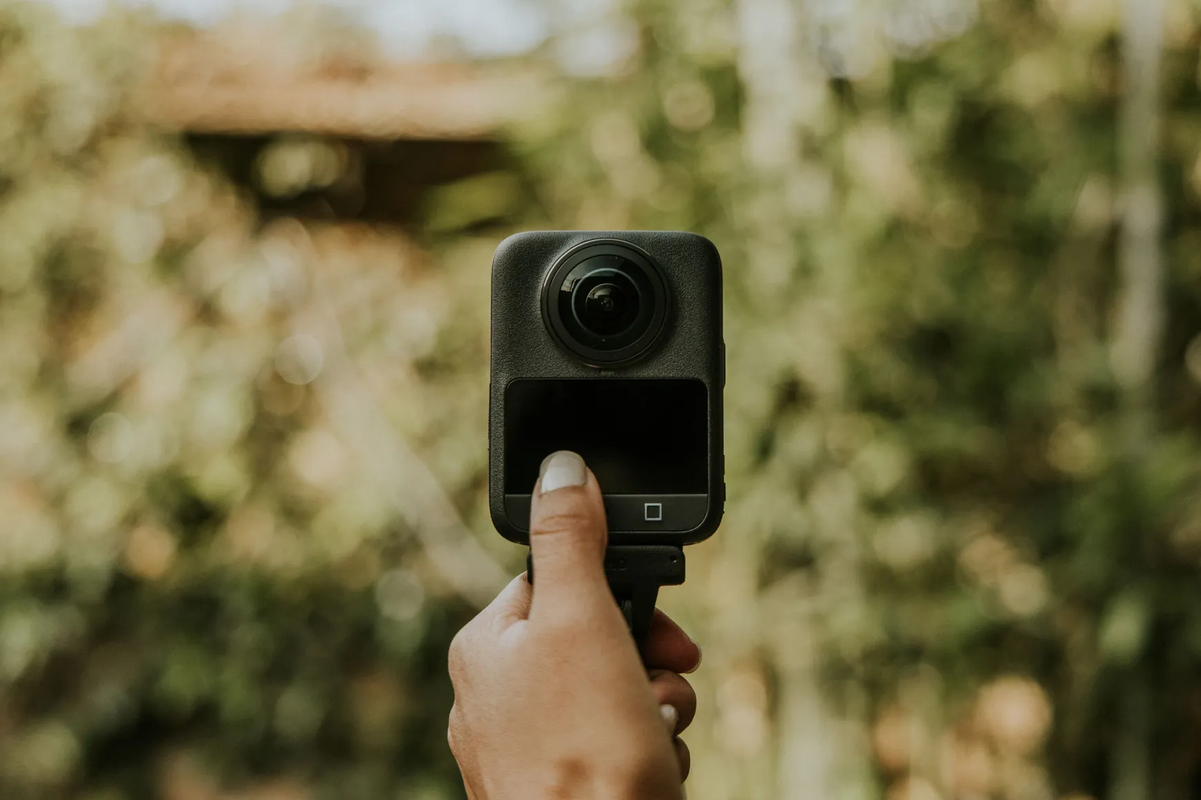Person holding a 360 camera outdoors with blurred green background.